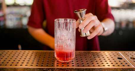 Cocktail making. Bar service. Bartender using jigger pouring red liquid to glass. Refreshing drink with ice cubes on metal counter.