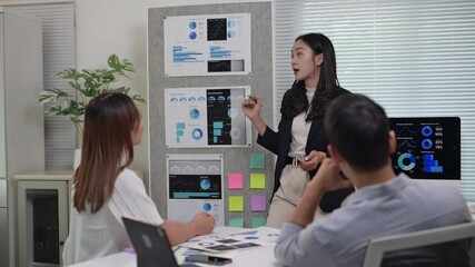 A woman is giving a presentation in front of a group of people. There are three people in the room, one of whom is looking at the woman