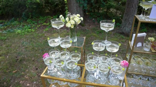 Beautifully arranged champagne glasses sparkle in the sunlight amidst lush greenery. The elegant setup, featuring pink and white flowers, creates a perfect wedding atmosphere.