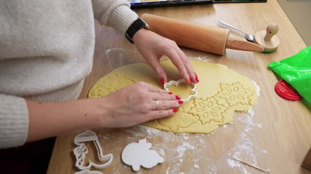 A close-up shot of hands pressing cookie cutters into dough, creating festive shapes. The scene captures warm natural lighting on a wooden table with baking tools nearby.