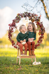 Lachende Geschwister sitzen gemeinsam auf einer Leiter in herbstlicher Kulisse mit K&uuml;rbissen und Bl&auml;tterkranz
