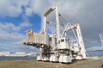 Large white self-propelled boarding ramp or mobile gangway parked at a port. This hydraulic passenger bridge connects cruise ships to the dock. Industrial machinery with wheels under a cloudy sky.