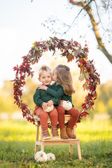 Liebevolles Geschwisterportr&auml;t im Herbst mit Kuss auf die Wange vor Bl&auml;tterkranz und K&uuml;rbissen