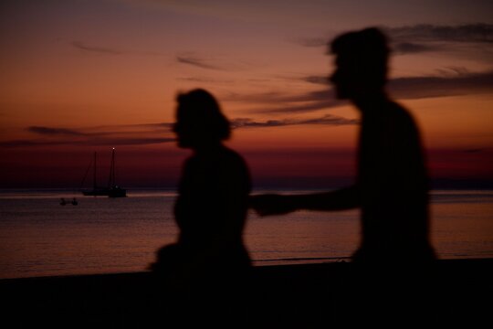 Silhouette di persone sfuocate passando sulla spiaggia al tramonto con il mare dietro. Colori del cielo arancioni.