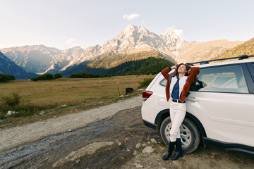 Naklejka premium Woman, car, mountains, road, landscape, travel, nature, relaxation. Young woman leaning against a white SUV on a gravel roadside with towering peaks and sweeping valley views outdoors.