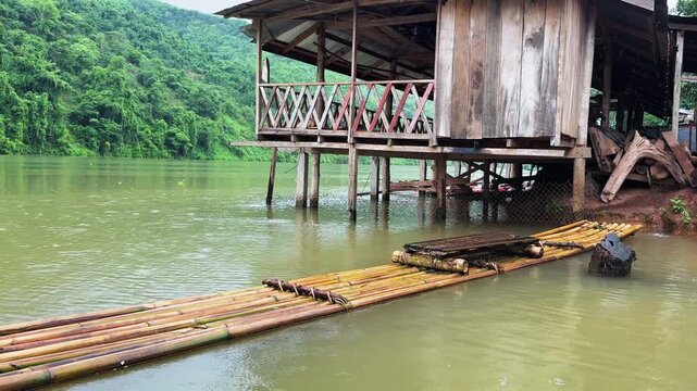 Rainy day on the Nam Ou River in Laos, with a wooden stilt house and bamboo raft moored on calm water below lush green hills.