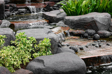 A detailed view of a multi-tiered garden water feature, featuring water cascading over and around large, dark gray rocks. © Ink N Propeller