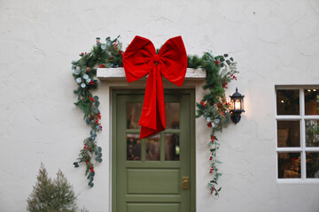 Fototapeta premium Merry Christmas decoration with large red bow and green garland over front door of white house exterior creating festive and welcoming holiday atmosphere for winter celebration season