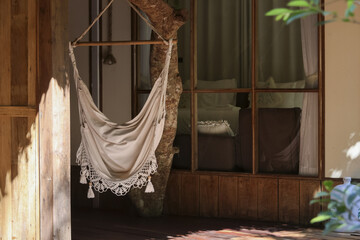 Relaxing beige cotton hammock chair hanging on wooden porch of rustic bungalow offering calm and...