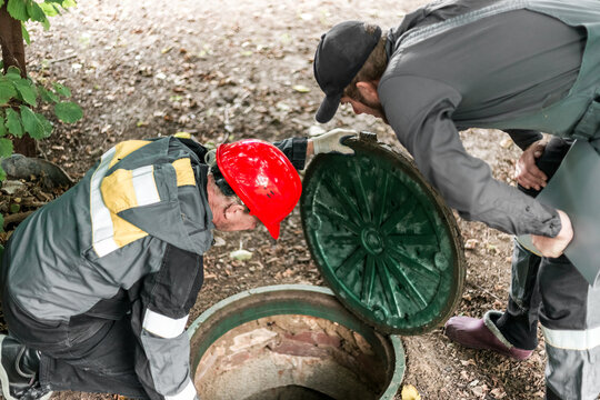 Male workers in overalls peer into an open manhole, checking utilities. Cleaning septic tanks and cesspools