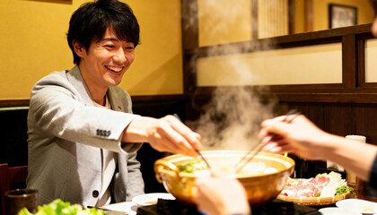 Smiling Asian man eating hot pot with a colleague at a restaurant. Friends enjoying a traditional Japanese nabe dinner