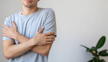A man in a light blue t-shirt with his arms crossed, conveying a sense of mental health and calmness in a minimalist indoor setting with a plant.