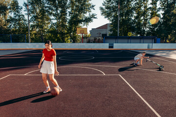 A young Hispanic girl dribbles a basketball on an outdoor court. A boy in the background rides a scooter. The scene is set in a sunny park with trees.
