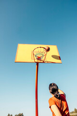 Unrecognizable teen girl throwing basketball toward hoop on outdoor court under clear blue sky, dynamic upward angle with ball nearing basket, youth sport, motivation, motion blur