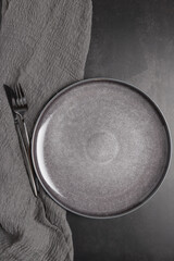 Vertical flat lay of an empty gray plate with black cutlery and linen fabric on dark background. Minimal and modern composition with copy space, suitable for design and decoration. 