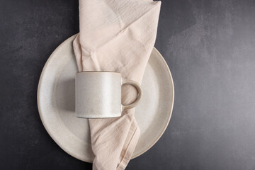 Flat lay view of beige ceramic mug and plate with linen napkin on dark textured background, clean and elegant composition. Horizontal image with copy space. 