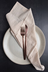 Vertical flat lay of ceramic plate with copper cutlery and neutral textile napkin on dark background.  Concept of modern table styling