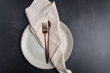 Horizontal flat lay of light ceramic plate with copper cutlery and beige linen napkin on dark gray background. Copy space.  Concept of minimalist table setting
