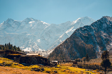 A horizontal landscape view of snow-covered high mountains in winter in Pakistan. Dry trees and a house on a hillside are visible on a clear, sunny day. Idea for a nature background with copy space.