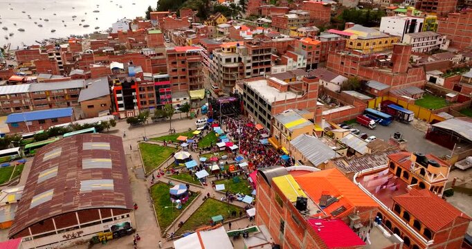 Aerial Orbital view of Carnival celebrations in Copacabana, Bolivia, showing crowds, market stalls and music stages in a dense urban landscape during a traditional South American festival