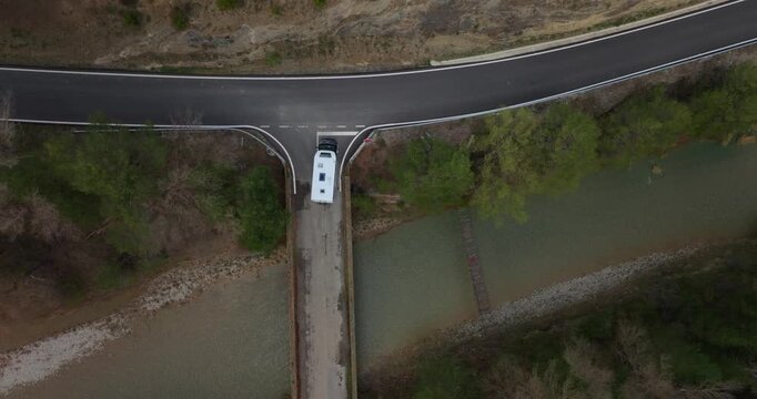 Top-down perspective of white integrated motorhome at a T-junction stop sign, crossing a narrow concrete bridge over a braided riverbed, navigating technical mountain passes