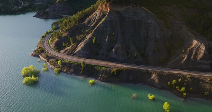 Aerial tracking shot of cyclists traversing the submerged ruins of the Antiguo Balneario de Tiermas and the arched masonry bridge of the A-1601 at the Yesa Reservoir, Spain.