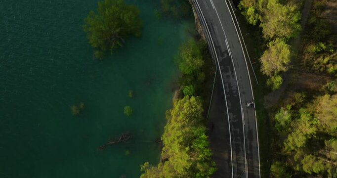 Mesmerizing top-down drone shot follows cyclists crossing the turquoise expanse of the Embalse de Yesa near the historic ruins of Tiermas, where the Pyrenees meet the water's edge, Aragon, Spain.