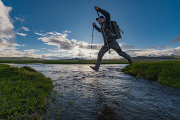 A hiker with trekking poles and a backpack crosses a mountain stream during golden hour. Lush green banks frame the figure moving right to left toward a meadow and peaks.