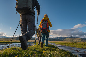 Hikers in bright gear with trekking poles walk along a mountain stream on an alpine meadow during golden hour. Rear medium shot captures them against a backdrop of peaks.
