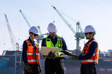 group of industrial engineers workers in a refinery - oil and gas processing equipment and machinery, engineers collaborate with a laptop, blueprint, and digital tablet at the oil storage tanks site.