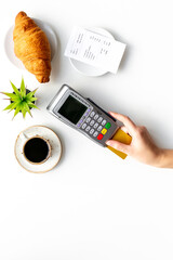 Hand is paying the bill by pos terminal in cafe or restaurant. Cup of black coffee and croissant, payment wireless machine and bill on white background, top view