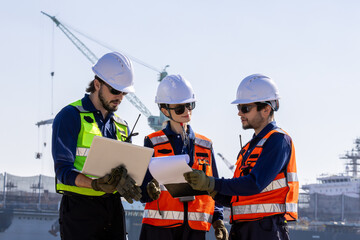 group of industrial engineers workers in a refinery - oil and gas processing equipment and machinery, engineers collaborate with a laptop, blueprint, and digital tablet at the oil storage tanks site.