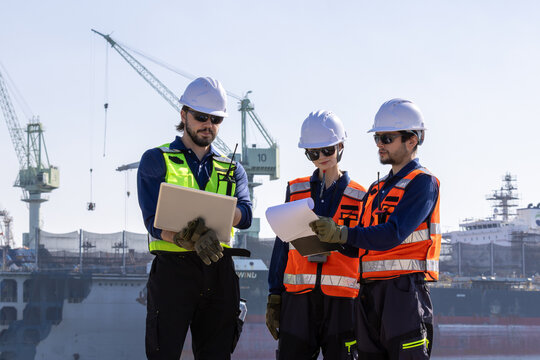 group of industrial engineers workers in a refinery - oil and gas processing equipment and machinery, engineers collaborate with a laptop, blueprint, and digital tablet at the oil storage tanks site. - Powered by Adobe