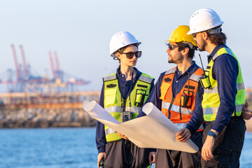 group of industrial engineers workers in a refinery - oil and gas processing equipment and machinery, engineers collaborate with a laptop, blueprint, and digital tablet at the oil storage tanks site.