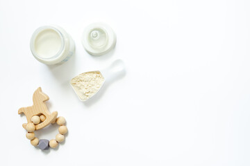 Powdered infant formula in jar lid and scoop with baby milk bottle and wooden teether on white background. Top view.