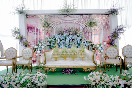 Elegant Javanese wedding stage with white sofas, blue and purple flowers, and Janur decorations.