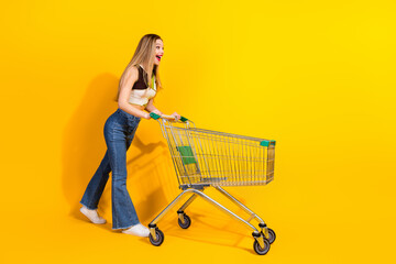 Cheerful young woman with a shopping cart against a vibrant yellow background, showcasing casual...