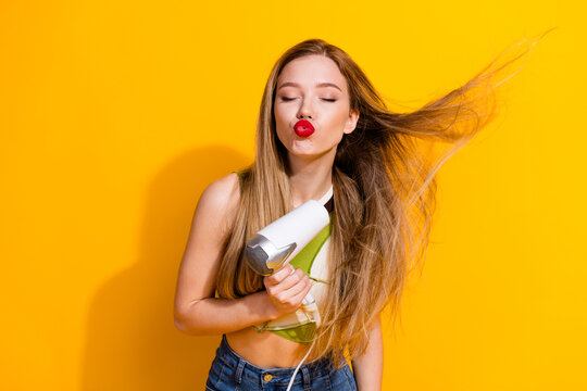 Playful young woman with long blonde hair blowing in the wind and a hairdryer, posing in front of a vibrant yellow background