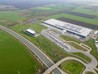 Aerial view of a large logistics warehouse with loading docks and trucks, distribution center surrounded by farmland. Supply chain and transportation concept