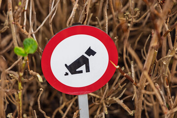 A red and white circular prohibition sign in a bush warning dog owners not to let their pets defecate in this specific area or garden.