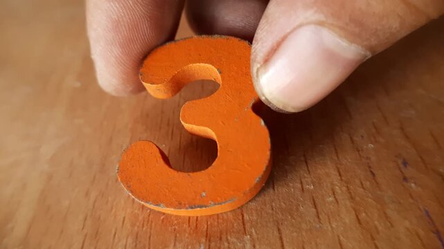 Hand holding an orange wooden number three toy on a light brown wood table surface background