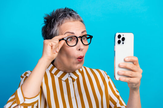 Aged female senior model with short gray hair wearing a striped shirt takes a selfie on a bright blue background