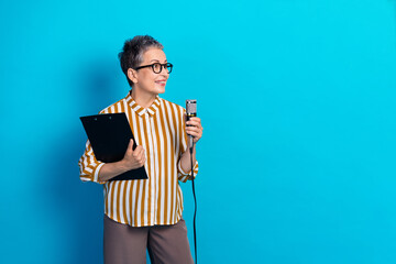 Senior woman with striped shirt holding clipboard and microphone stands in bright blue studio ready...