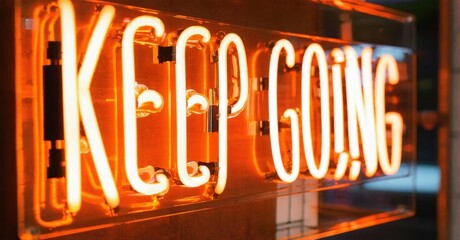 Bright neon sign displays KEEP GOING. Warm orange glow illuminates lettering against dark background. Inspiring message, simple design.