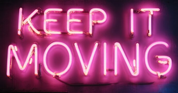 Bright pink neon sign displays motivational phrase keep it moving. Dark background emphasizes glowing text. Simple, direct message.