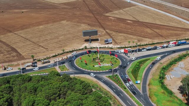 Aerial view of a busy roundabout outside Casablanca, surrounded by fields and traffic, with red flags and a large central billboard.