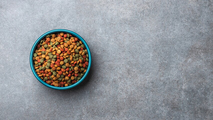 A blue bowl filled colorful dry pet food kibble on gray stone textured background. © PixelArt_25