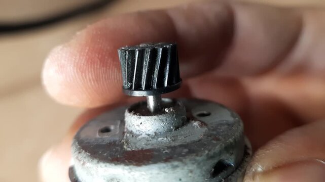 Human fingers holding a small dynamo motor with a black plastic gear on a rusty metal shaft in a macro shot