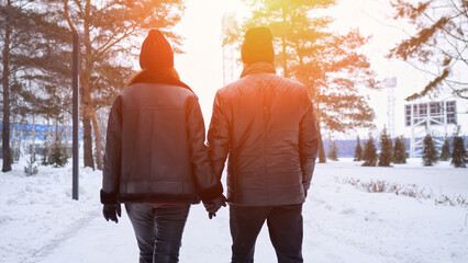 Couple Holding Hands Walking Through Snowy Park, Sunset Backlight Creates Warm Glow, Tall Pines Line Path, Bundled Jackets And Beanies, Peaceful Winter Atmosphere, Intimate Connection And Gentle Pace