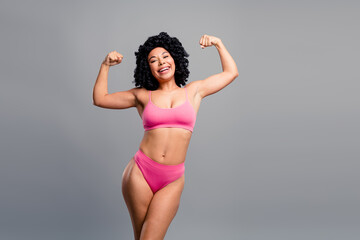 Confident young woman in pink bikini flexes her arms  smiles fitness lifestyle commercial image in studio with grey background promoting beauty health and body positivity
